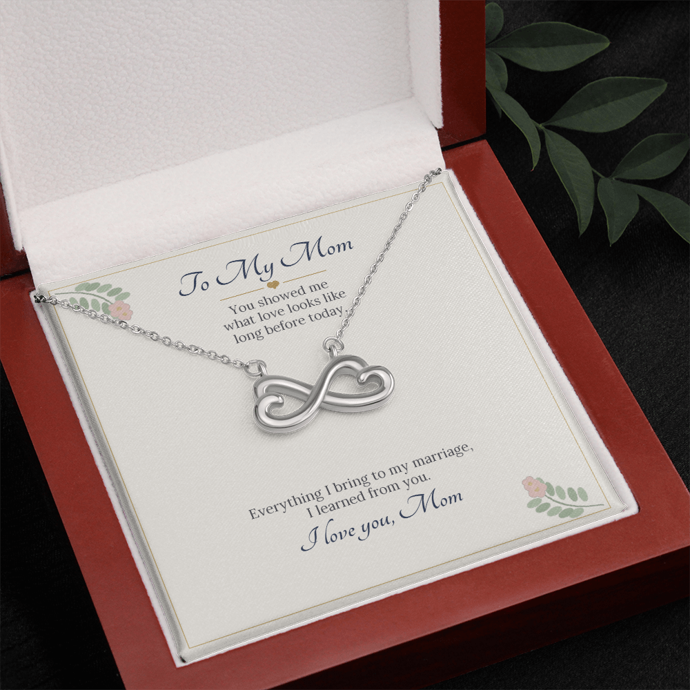 White gold finish infinity heart necklace on the To My Mom wedding card in an open wooden luxury box beside green leaves