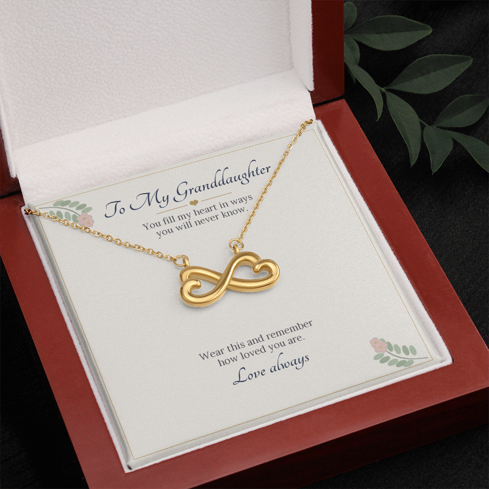 Yellow gold finish infinity heart necklace on the To My Granddaughter card in an open wooden luxury box, seen from above with leaves beside it